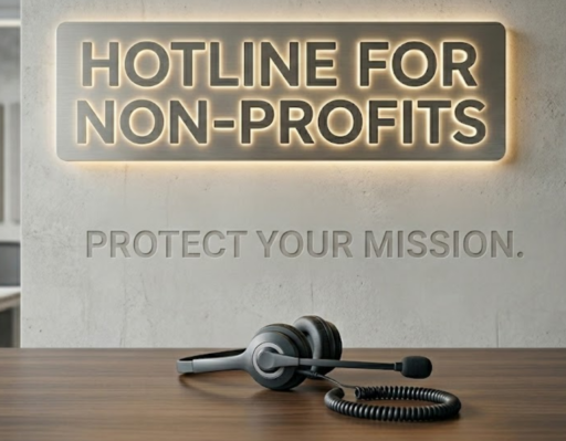 A professional photo of an office reception desk features a modern headset resting on the polished wooden surface, with a coiled cord trailing nearby. On the concrete wall behind the desk, a large, illuminated sign made of metal with glowing letters proudly displays the text "HOTLINE FOR NON-PROFITS." Directly below the sign, the phrase "PROTECT YOUR MISSION." is subtly debossed into the wall.